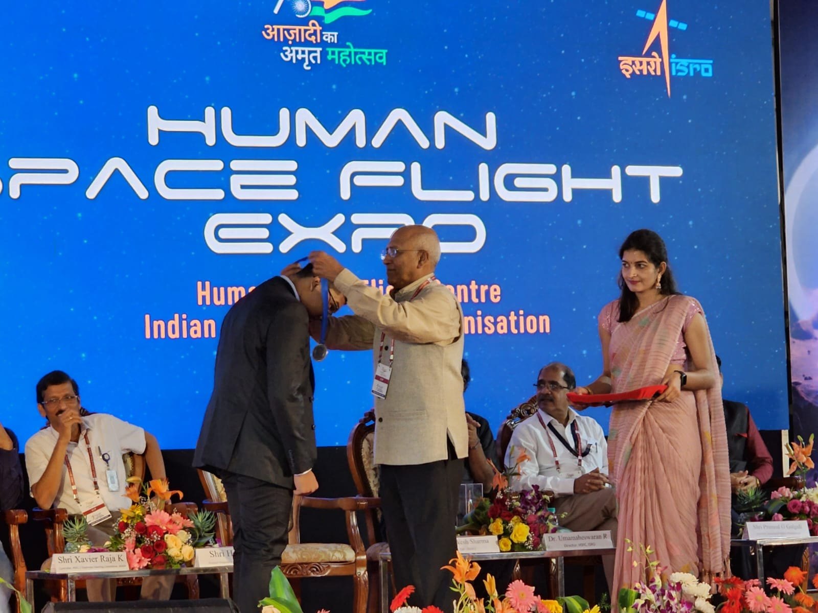 Rakesh Sharma placing a medal around Senaajit's neck on stage at ISRO's Human Space Flight Expo